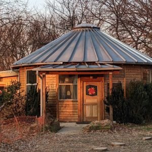Exterior view of The Carson octagonal kit home featuring wood siding, metal roof, front porch, and compact footprint surrounded by trees.
